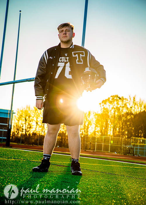 A young male football player wearing a varsity jacket and uniform stands on a football field holding a helmet during his Troy senior pictures session, with goalposts and the sun setting in the background.
