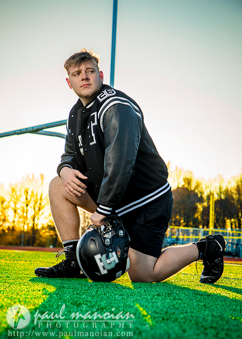 A young male athlete in a letterman jacket kneels on a football field at sunset during his Troy senior pictures session, holding a helmet. Goalposts and trees are visible in the background.