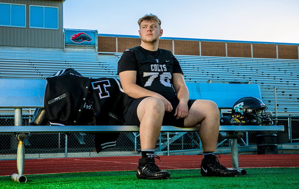 A high school senior football player in a black uniform sits on a bench in a stadium during his Troy senior pictures session. His helmet and letterman jacket rest beside him, with empty bleachers in the background.