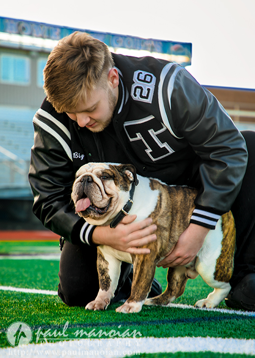 A young man in a varsity jacket kneels on a football field during his Troy senior pictures session, gently hugging a smiling English bulldog. The stadium seats are visible in the background, and both appear happy and relaxed.