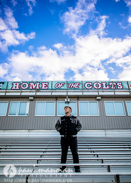 A person in a letterman jacket stands on empty bleachers beneath a sign reading "HOME OF THE COLTS," with a bright blue sky and scattered clouds—a classic moment from a Troy senior pictures session.