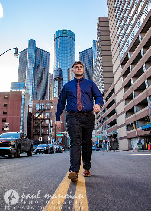 A man in business attire confidently walks down the middle of a city street, with tall modern buildings and cars in the background under a blue sky—a striking Troy senior pictures session photographed by Paul Manoian.