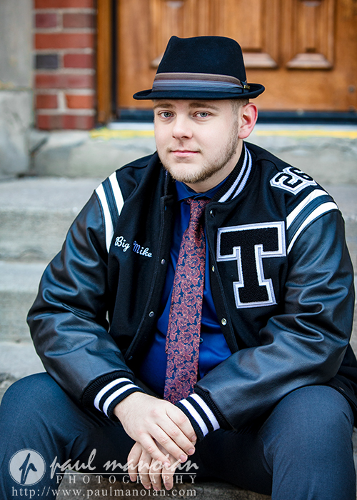 A man wearing a black fedora, a letterman jacket with a "T" and "Big Mike" on it, a dress shirt, and patterned tie sits on outdoor steps in front of a wooden door—capturing his Troy senior pictures session.