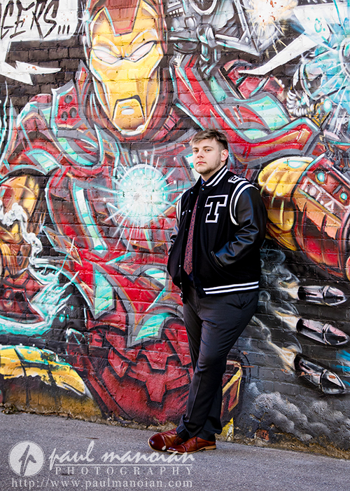 A young man in a letterman jacket leans against a brick wall with colorful Iron Man graffiti during his Troy senior pictures session. He stands with one leg crossed, hands in pockets, and looks thoughtfully into the distance.