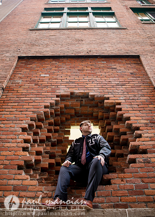 A young man in a letterman jacket sits in a circular hole in a brick wall, looking to the side. Tall building windows rise above him. Photography credit appears at the bottom, highlighting this Troy senior pictures session.