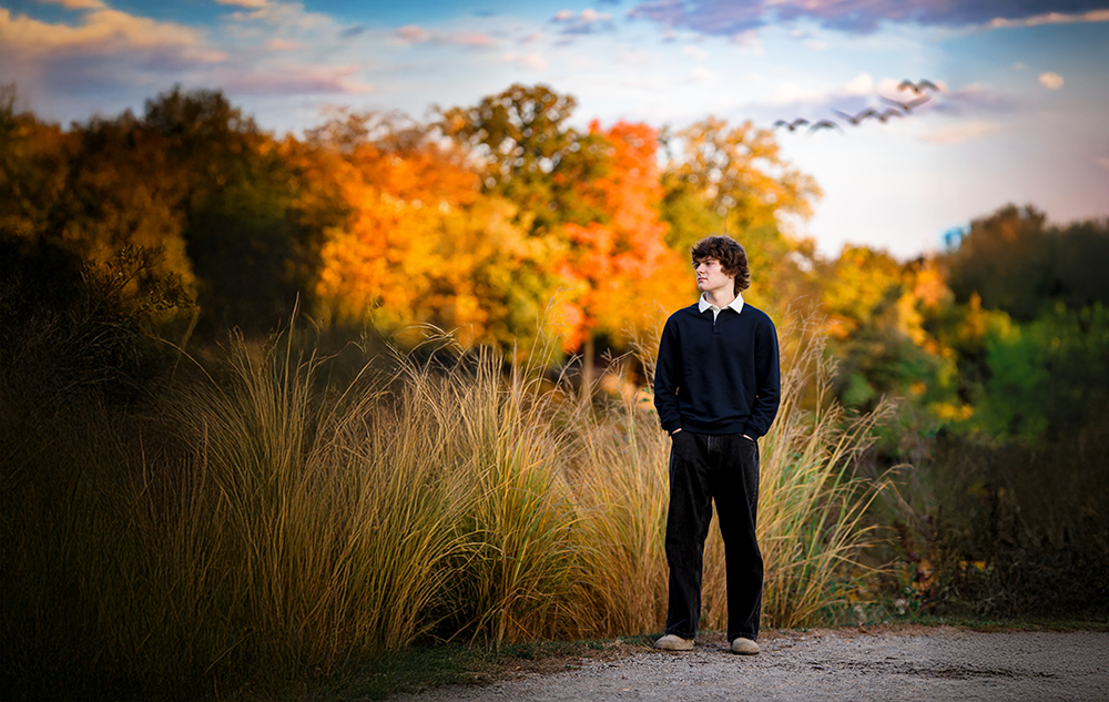 A high school senior boy stands on a path surrounded by tall grass during his fall senior pictures session, with colorful autumn trees and a flock of birds flying in the sky behind him. He looks to his left, dressed in a dark sweater and pants.