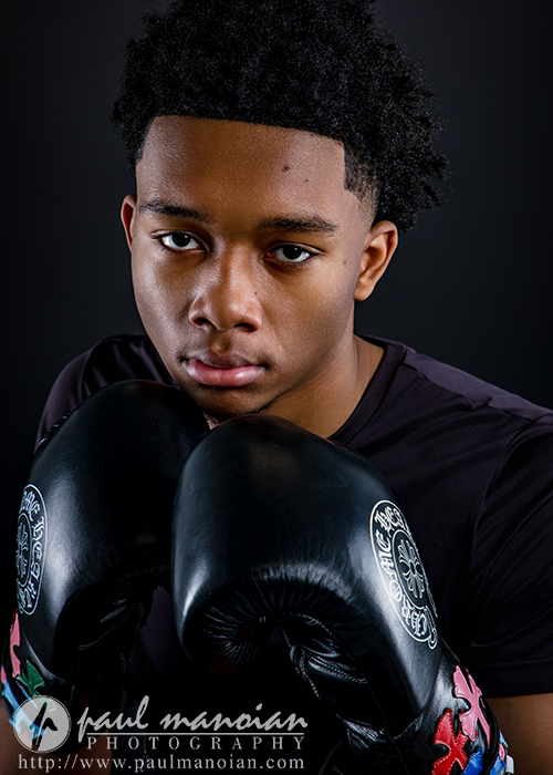 A young man with short curly hair wearing black boxing gloves and a black shirt poses against a dark background, looking directly at the camera with a serious expression—perfect for striking boxing senior pictures. The gloves have white and colorful designs.