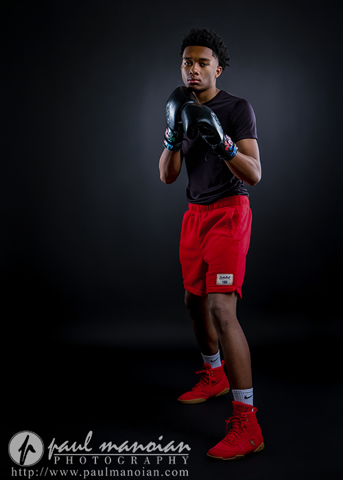 A young male boxer stands in a fighting stance, wearing black gloves, a black t-shirt, red shorts, and red boxing shoes against a dark background—perfect inspiration for dynamic boxing senior pictures.