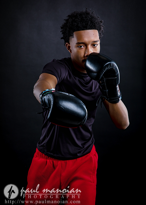 A young man wearing black boxing gloves, a black t-shirt, and red shorts poses in a boxing stance against a dark background—perfect for dynamic boxing senior pictures as he extends one arm forward as if throwing a punch.