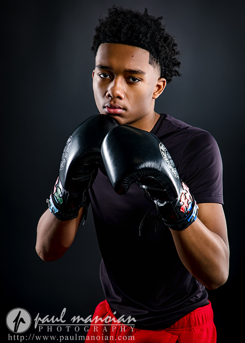 A young man wearing black boxing gloves and a black T-shirt stands in a boxing stance, looking forward against a dark background—perfect for dynamic boxing senior pictures. Photography watermark and website appear at the bottom.