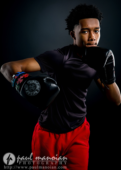 A young male boxer wearing black gloves, a black shirt, and red shorts poses in a fighting stance against a dark background—perfect for unique boxing senior pictures. The image is professionally lit and features a photography watermark at the bottom.