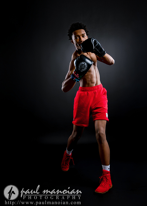 A young male boxer in red shorts and red shoes poses in a boxing stance with raised gloves against a black background, perfect for dynamic boxing senior pictures. The lighting highlights his muscular physique.