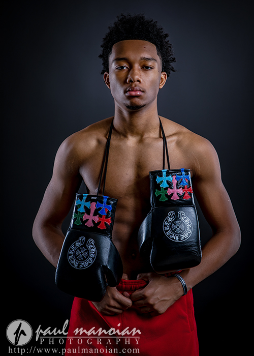 A young man stands against a dark background, shirtless and wearing red shorts. With black boxing gloves decorated with colorful stars hanging around his neck, he confidently poses for his boxing senior pictures.