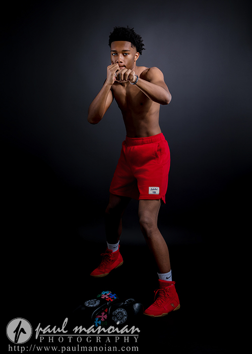 A young man stands in a boxing stance, wearing red shorts and red sneakers, against a dark background. Perfect for boxing senior pictures, he is shirtless and facing forward, with boxing gloves placed on the floor in front of him.