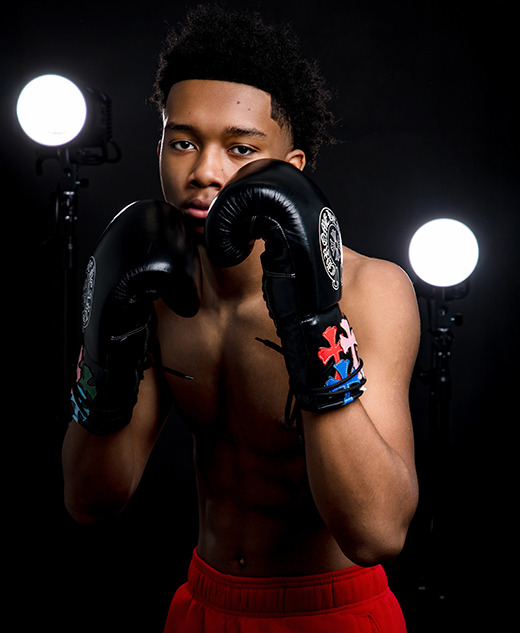 A young male boxer with short curly hair poses in a boxing stance, wearing black gloves and red shorts—perfect for dynamic boxing senior pictures—framed by two bright studio lights against a dark background.
