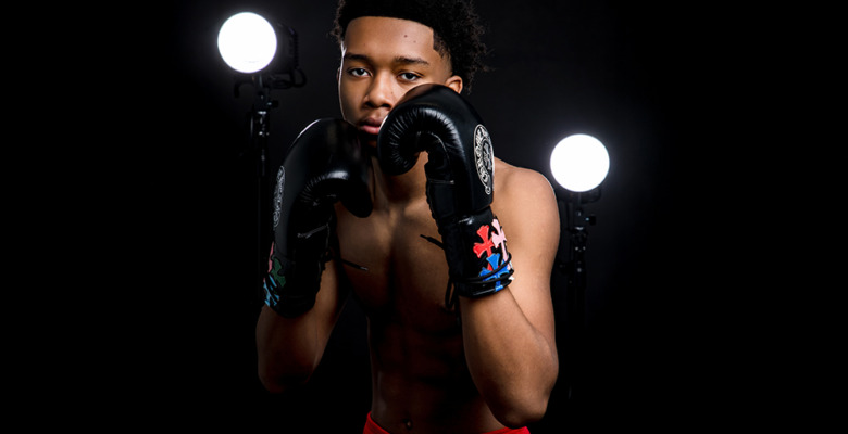 A young male boxer with short curly hair poses in a boxing stance, wearing black gloves and red shorts—perfect for dynamic boxing senior pictures—framed by two bright studio lights against a dark background.
