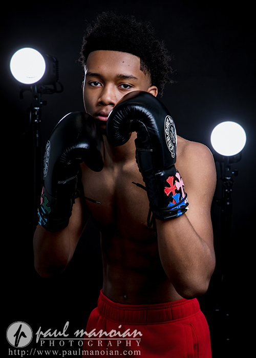 A young male boxer in black gloves and red shorts poses with fists raised for his boxing senior pictures in a dark studio, illuminated by two bright lights behind him. The logo "paul manoian photography" appears at the bottom of the image.