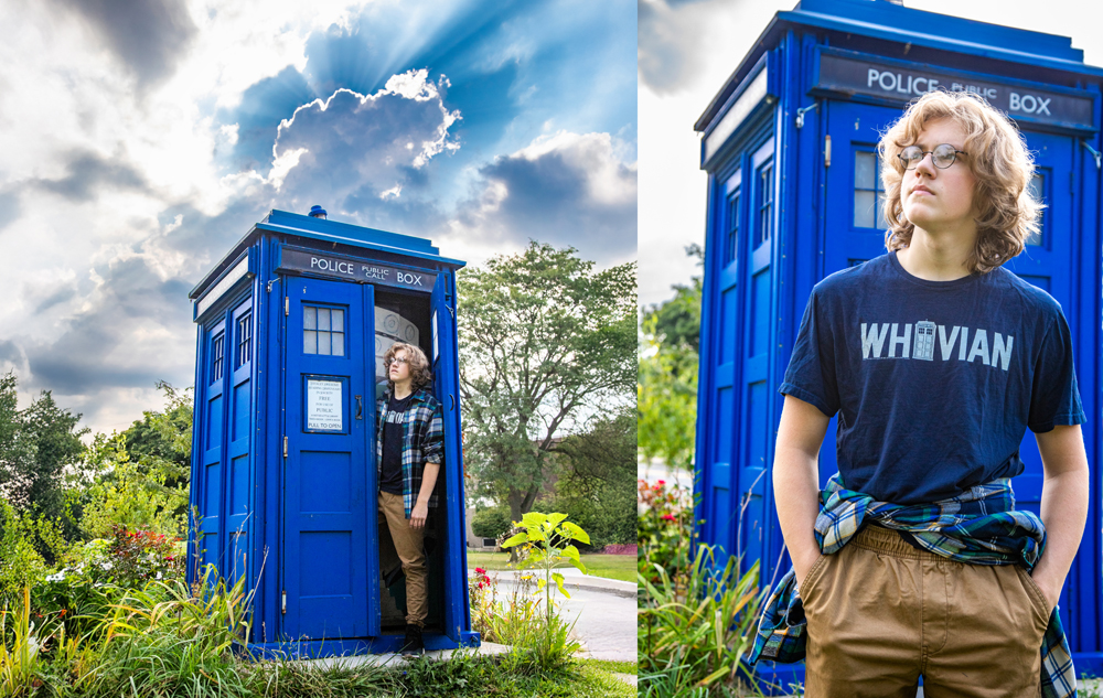 A high school senior boy with shoulder-length blond hair stands by a bright blue police box resembling the TARDIS from Doctor Who, wearing glasses, a "WHOVIAN" T-shirt, khaki pants, and a flannel shirt tied at the waist during a summer senior pictures session.