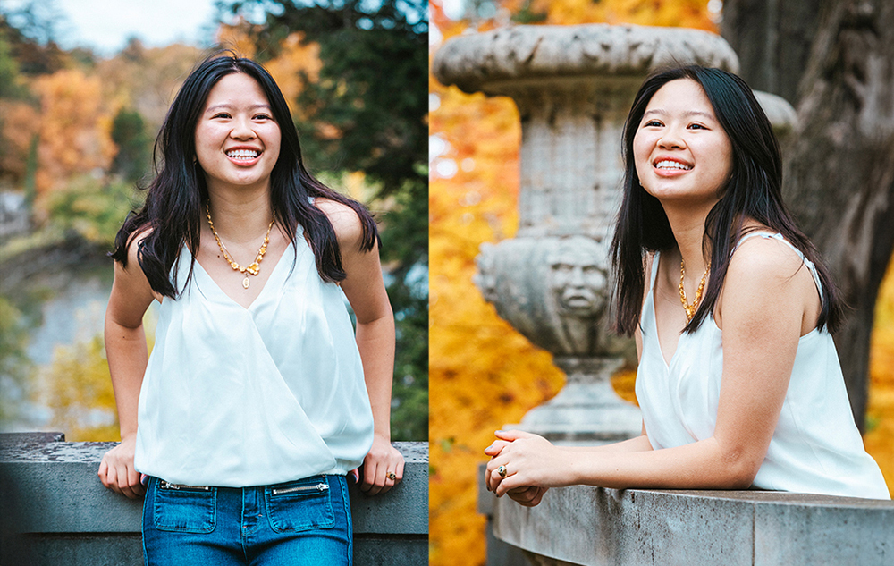 A high school senior girl with long dark hair wearing a white sleeveless top and blue jeans smiles while leaning on a stone railing outdoors during her fall senior pictures session, with autumn trees and a stone urn in the background.