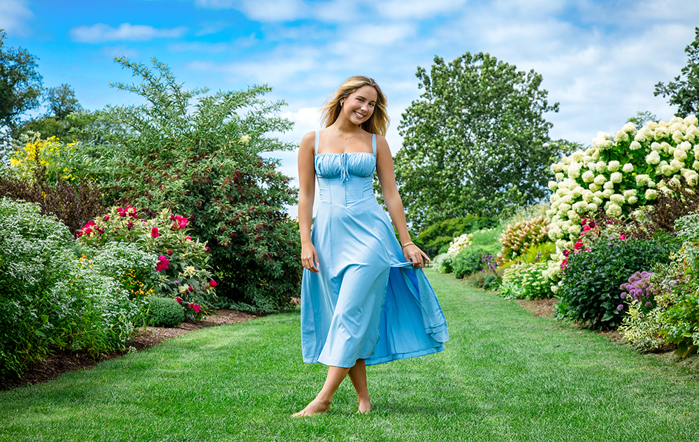 A high school senior girl in a light blue dress stands barefoot on a green lawn, smiling and holding her dress during a summer senior pictures session. She is surrounded by colorful flowers and lush greenery under a partly cloudy sky.