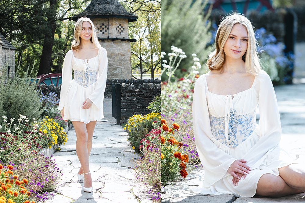 A high school senior girl in a white dress with blue embroidery stands and sits among colorful flowers on a stone path during her summer senior pictures session, with a rustic stone building and lush greenery in the background, smiling at the camera.