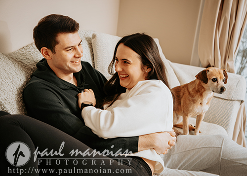 A smiling couple sits on a couch, cuddling and laughing together during their engagement portrait session. A small brown and white dog stands beside them on the couch, looking toward the camera. Warm, natural light fills the cozy living room.