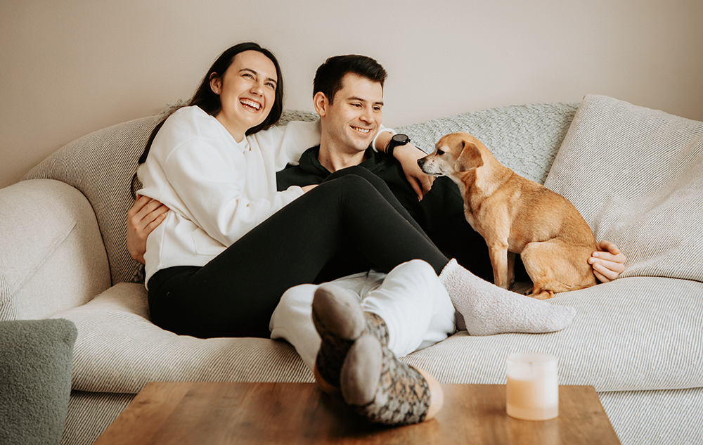 A smiling couple sits closely together on a beige couch, with a small brown dog beside them. The woman laughs while leaning on the man, and a lit candle is on the wooden table in the foreground.