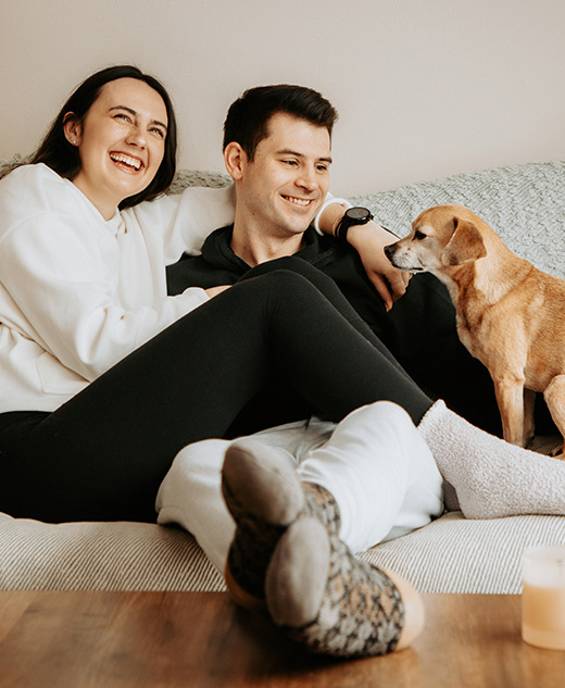 A woman and man sit closely together, smiling and relaxing on a couch during their engagement portrait session. A small brown dog sits next to them, looking at the man, while a lit candle rests on the wooden table in front of them.