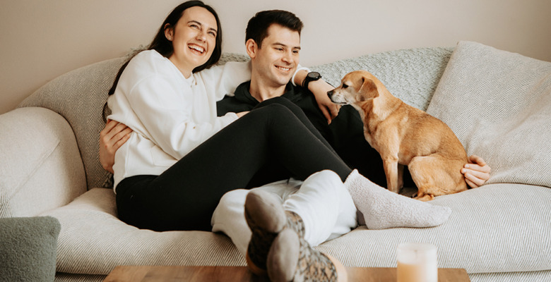 A woman and man sit closely together, smiling and relaxing on a couch during their engagement portrait session. A small brown dog sits next to them, looking at the man, while a lit candle rests on the wooden table in front of them.