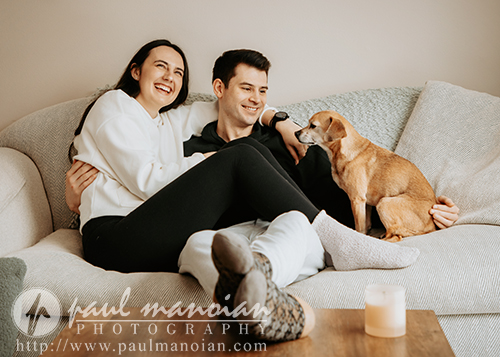 A smiling couple sits closely together on a beige couch during their engagement portrait session, with a small brown dog beside them looking at the man. The warm, relaxed atmosphere is enhanced by a candle on the table in the foreground.