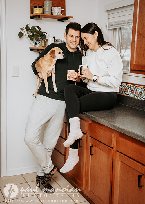 A smiling couple relax in a cozy kitchen during their engagement portrait session, with the man holding a small brown dog and the woman sitting on the counter holding a mug. Shelves with jars and dishes are visible in the background.