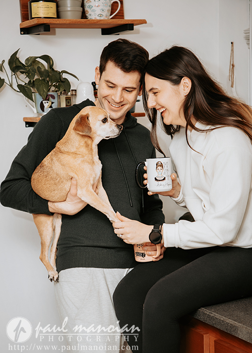 A smiling couple in a kitchen holds a small brown dog during their engagement portrait session. The man gently supports the dog while the woman, seated with a mug, touches its paw. They appear happy and relaxed together.