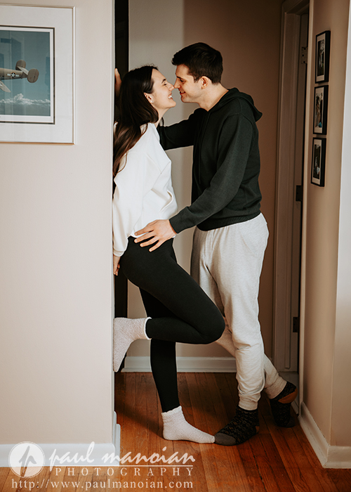 A young couple stands in a hallway during their engagement portrait session, smiling at each other. The woman leans against a doorframe with one knee bent as the man gently touches her waist, both appearing happy and relaxed in a cozy home setting.
