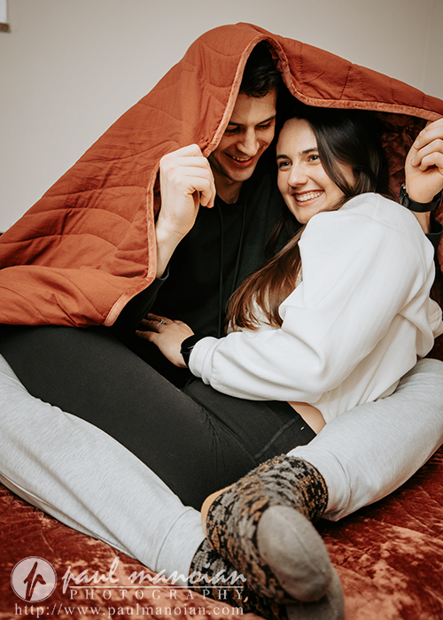 A smiling couple sits closely together under a brown blanket, creating a cozy tent. Captured during their engagement portrait session, they sit on a cushioned surface looking happy and comfortable. The photo includes a watermark for Paul Mannion Photography.