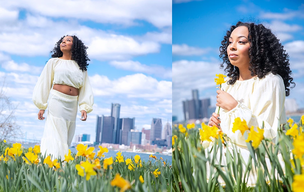 A woman with curly hair in a light yellow outfit walks and poses among blooming yellow daffodils, capturing beautiful spring senior pictures in Detroit with the city skyline and blue sky with clouds in the background.