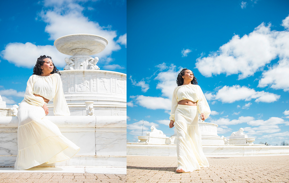 A high school senior girl in a flowing white outfit stands confidently by a white marble fountain under a bright blue sky with scattered clouds, posing with poise—an ideal scene for a spring senior pictures session. Her dark hair is styled in loose waves.