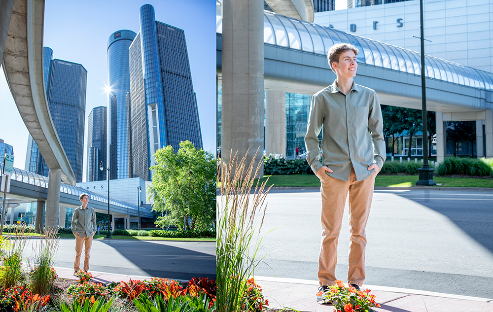 A high school senior boy in a light green shirt and tan pants stands outdoors near flower beds, with tall modern skyscrapers and a covered walkway in the background on a sunny day during his spring senior pictures session.