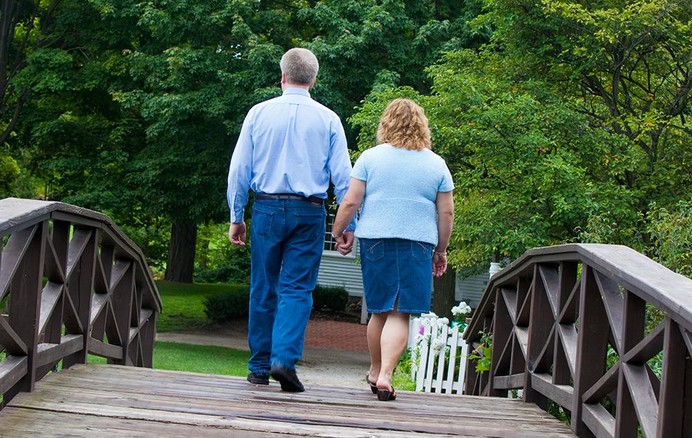 A man and a woman, both in blue, walk hand in hand across a wooden footbridge in a lush park. Captured by a wedding engagement portraits photographer, their backs face the camera as trees and a white gate frame the serene moment.