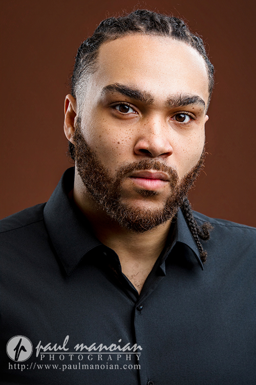 A man with braided hair and a trimmed beard wears a black button-up shirt, looking seriously at the camera. Set against a solid brown background, this Detroit actor headshot includes a watermark for Paul Manoian Photography.