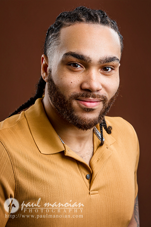 A man with long braids, a beard, and mustache, wearing a mustard yellow collared shirt, smiles softly at the camera against a brown background. The photo features the "paul manonian photography" watermark, capturing Detroit actor headshots style.