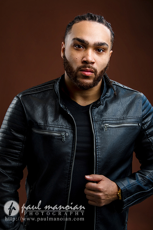 A man with braided hair and a beard wears a black leather jacket and black shirt, standing against a brown background. He looks confidently at the camera with one hand gripping his jacket, capturing classic Detroit actor headshots style. Photography credit appears at the bottom.