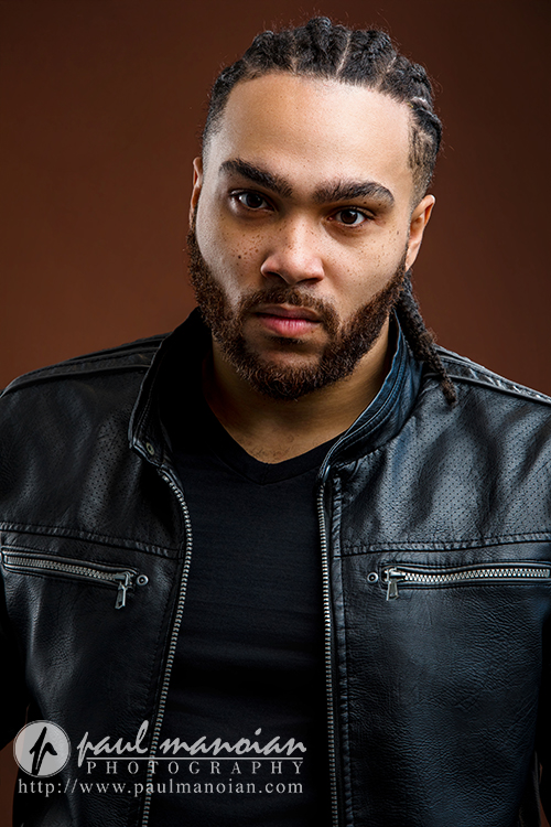 A man with braided hair and a beard wears a black leather jacket over a black shirt, looking seriously at the camera against a brown background—perfect for Detroit actor headshots. Photography credit is visible in the bottom left corner.