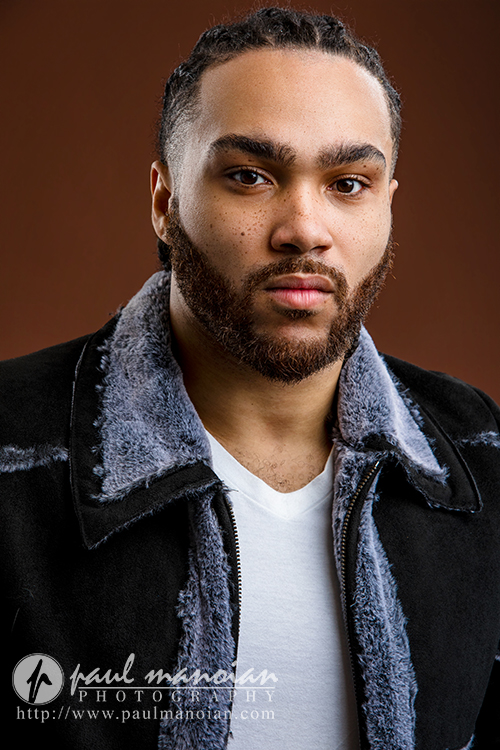 A man with braided hair, a full beard, and mustache wears a black and gray shearling jacket over a white shirt. Shot in the style of Detroit actor headshots, he stands against a brown background, looking directly at the camera with a neutral expression.