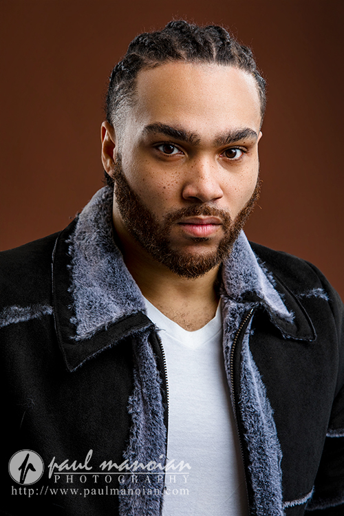A man with short braids, a trimmed beard, and mustache wears a black jacket with a gray furry collar over a white shirt, standing against a plain brown background and looking seriously at the camera—perfect for Detroit actor headshots.