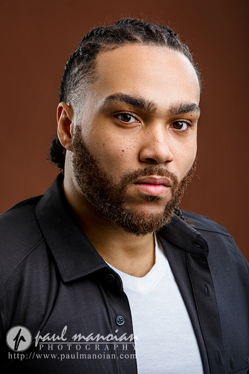 A man with braided hair and a trimmed beard wears a black shirt over a white T-shirt, looking seriously at the camera against a brown background—an example of Detroit actor headshots, with a watermark for Paul Manoian Photography.