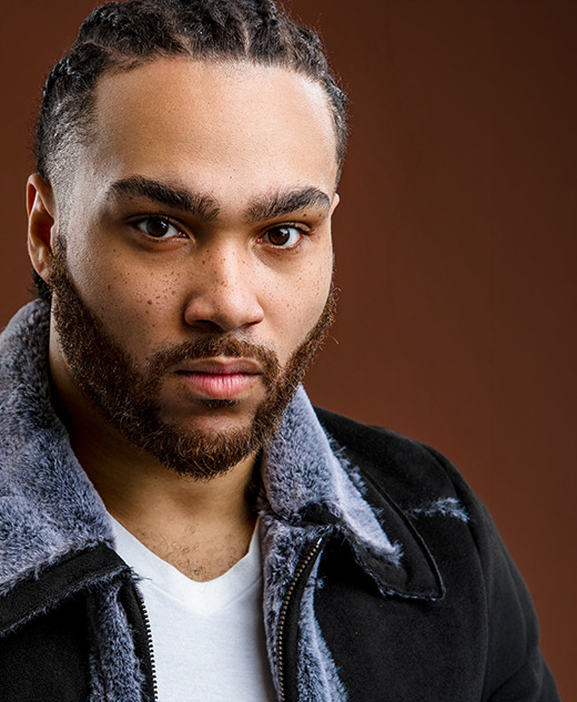 A man with braided hair and a beard, wearing a black shearling jacket over a white shirt, poses for a Detroit actor headshots session with award winning photographer, Paul Manoian.