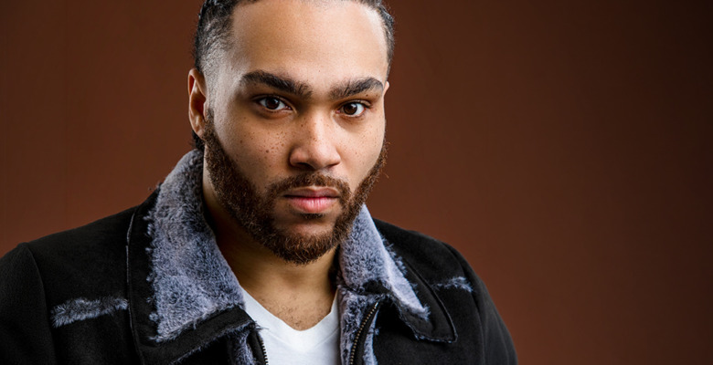 A man with braided hair and a beard, wearing a black shearling jacket over a white shirt, poses for a Ann Arbor actor headshots session with award winning photographer, Paul Manoian.