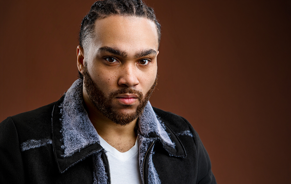 A man with braided hair and a beard, wearing a black shearling jacket over a white shirt, poses for a Detroit actor headshots session with award winning photographer, Paul Manoian.