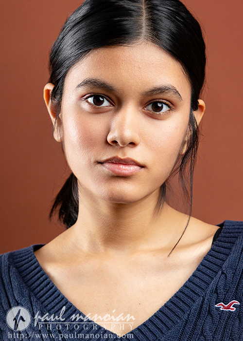 A young woman with long dark hair in a low ponytail wears a navy blue V-neck sweater. Captured by an Ann Arbor actor headshots photographer, she looks directly at the camera with a neutral expression against a warm brown background.