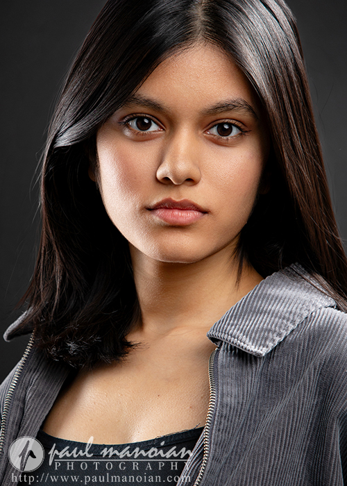 A young woman with straight dark hair and brown eyes looks directly at the camera. She is wearing a gray collared jacket over a black top. Captured by an Ann Arbor Actor Headshots Photographer, the dark background creates a professional studio portrait look.